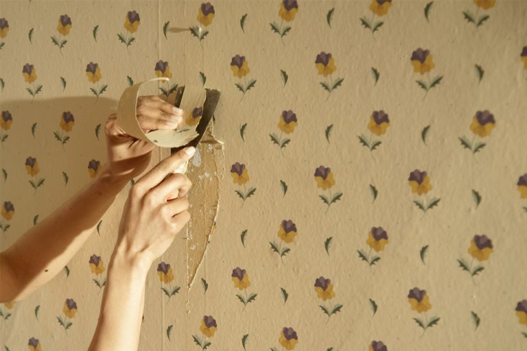 A hand scraping old wallpaper off the wall.